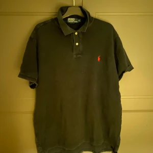 Svart långärmad pikétröja från Ralph Lauren - Säljer en klassisk svart långärmad pikétröja från Ralph Lauren med den ikoniska loggan broderad i rött på bröstet. Tröjan har en krage med knappar och är perfekt för en stilren look.