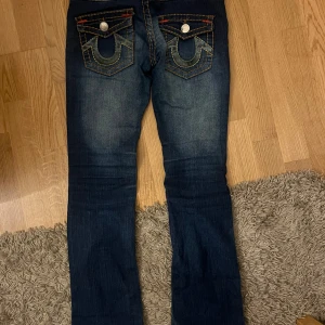 Mörkblå jeans från True Religion - Snygga mörkblå jeans från True Religion med ljusa sömmar och ikoniska bakfickor med knappar. Jeansen är lågmidjade. Midjemåttet är 38 rakt över och innerbens längden är 77. Säljer då det för små för mig 
