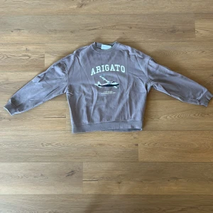 Grå/lila sweatshirt med tryck - Säljer en grå/lila Axel Arigato sweatshirt med texten 'ARIGATO' och ett flygplanstryck på framsidan. Tröjan har långa ärmar och ribbade muddar vid ärmslut och nederkant. Perfekt för en avslappnad stil.