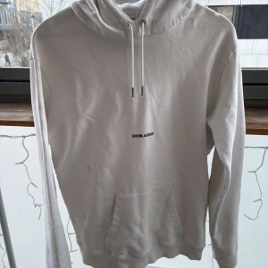 Vit hoodie från Saint Laurent - Stilren vit hoodie från Saint Laurent med justerbar huva, perfekt för både vardagsbruk och casual elegans. 💯 Äkta och verifierad av Sicuro, vilket garanterar autenticitet. Storlek M men kan även passa en S beroende på önskad passform (oversized eller regular). Materialet är mjukt och slitstarkt, vilket gör den både bekväm och långvarig.