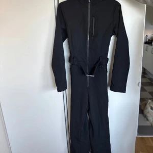 Svart skisuit  - En svart supersnygg skisuit från Zara. Köpt förra vintern och bara använd en skidsemester två dagar, tyvärr blivit för stor för mig denna säsong därav sälj. Jättefint skick, sitter snyggt på kroppen och är framförallt skön! Dragkedja framtill och justerbart bälte i midjan. Utrustad med Recco- reflektor för extra säkerhet. Snygga detaljer också. Inga revor från pjäxor eller dylikt finns. Liten i storlek, skulle vilja påstå att den är mer utav en M än en storlek L. Ordinarie pris 1399:- 