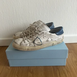 Vita sneakers från Philippe Model - Snygga vita sneakers från Philippe Model med blå detaljer och snörning. Skorna har en robust sula och en stilren design som passar perfekt för en avslappnad look. Dom är använda en hel del men finns fortfarde stor potential t.ex lämna in för att få sulan hel. Skick 6/10 och dom är i storlek 43 