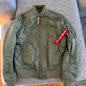 Grön bomberjacka från Alpha Industries - Säljer en grön bomberjacka från Alpha Industries i storlek medium. Jackan har en klassisk design med dragkedja och ribbade muddar. Den har en orange insida och en ikonisk röd tagg på ärmen. Perfekt för en cool och avslappnad stil.