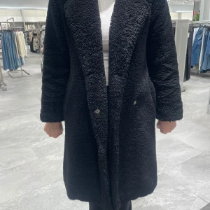 Svart teddyjacka - Säljer en mysig svart teddyjacka med långa ärmar och knappar framtill. Perfekt för kyliga dagar och ger en stilren look. Jackan är i en längre modell som håller dig varm och bekväm.