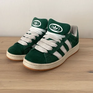 Gröna Adidas Campus sneakers - Snygga gröna Adidas Campus sneakers i mocka med vita detaljer och snörning. De har den klassiska tre-randiga designen på sidorna och en gummisula i ljusbrun färg Använd några fåtal gånger. 