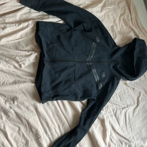 Nike tech fleece svart - Säljer en svart huvtröja från Nike med dragkedja och två fickor framtill i barnmodell. Tröjan har en enkel design med diskreta ränder över bröstet och en huva för extra komfort. Perfekt för en avslappnad stil. 