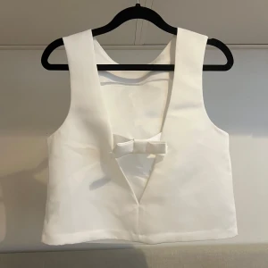 Vit topp från Divided - Säljer en stilren vit topp från Divided. Den har en söt rosett baktill och är ärmlös med en enkel design. Perfekt för att lägga till en touch av elegans till din outfit. Har en liten fläck men går att tvätta bort