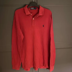Röd långärmad pikétröja från Ralph Lauren - Säljer en klassisk röd långärmad pikétröja från Ralph Lauren med krage och knappar. Perfekt för en stilren look. Tröjan har en broderad logotyp på bröstet.