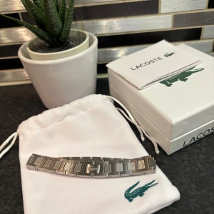 Silverfärgat armband från Lacoste - Elegant silverfärgat länk armband från Lacoste med ett stilrent och modernt designmönster. Armbandet har en justerbar passform och kommer i originalförpackning med en vit tygpåse för förvaring. Helt nytt! Hör av er vid eventuella frågor eller funderingar🤩