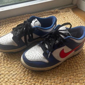 Nike Dunk sneakers med röd och svart swoosh - Snygga Nike Dunk sneakers i vitt, blått och svart med en röd och en svart swoosh. Det röda swooshet finns på båda skorna utåt och det svarta swooshet finns också på båda skorna fast inåt, som ni kan se på första och andra bilden.. Skorna har svart snörning och perforerad tå för extra ventilation. Perfekta för en sportig look. Använda men i gott skick.