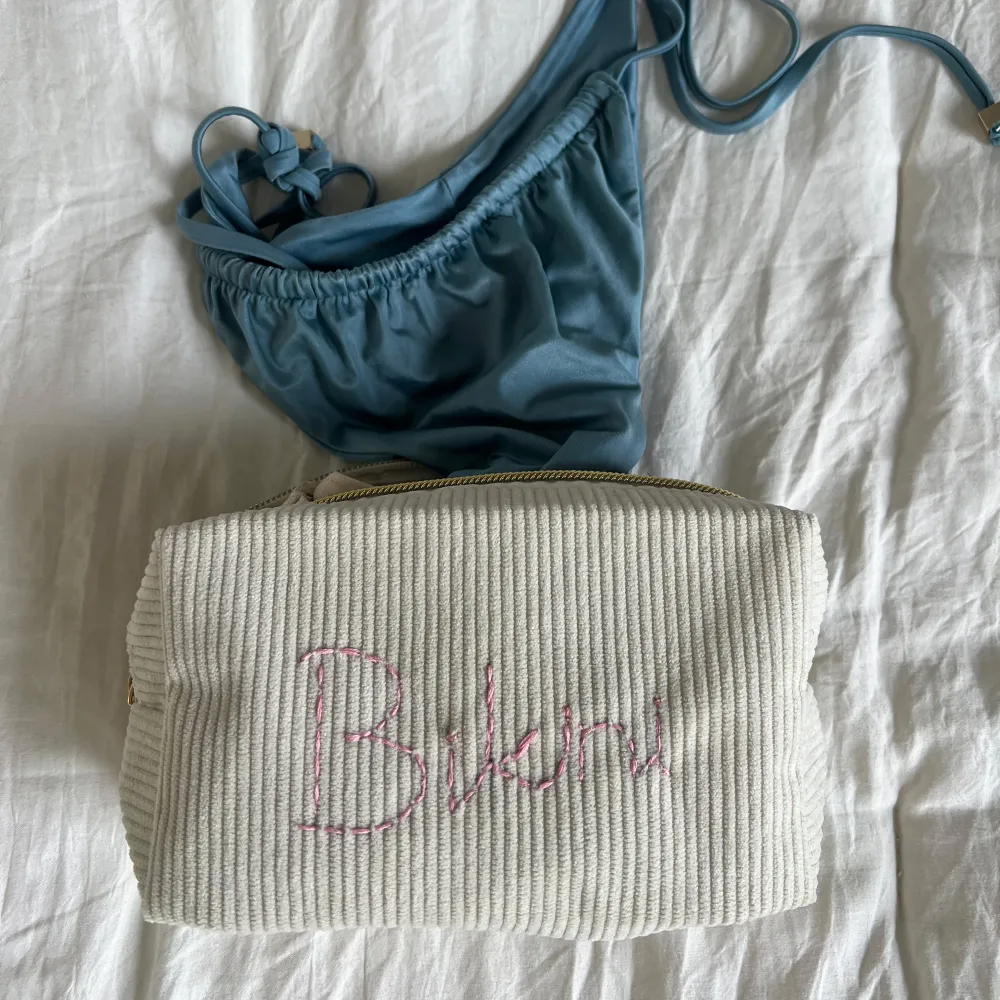 En vit ribbad förvaringspåse med texten 'Bikini' (går även att be om annan personlig broderad text) broderad i rosa. Smidig att ta med på resan! Storlek: 17x10x6cm. Laukut & Käsilaukut.