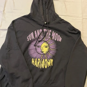 Svart hoodie med sol- och månmotiv - Säljer en svart hoodie med ett coolt tryck av solen och månen i lila och gult. Texten 'Sun and the Moon Harmony' pryder framsidan. Perfekt för en avslappnad stil.