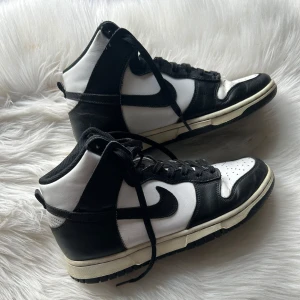 Svartvita Nike Dunk sneakers - Snygga svartvita Nike Dunk sneakers med hög design och klassisk snörning. Perfekta för en stilren look med sin kontrast mellan svart och vitt. Skorna har en bekväm sula och ikonisk Nike-logga på sidan.