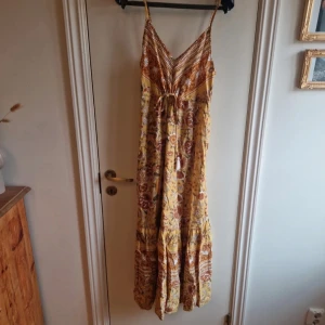 Gul blommig långklänning - Säljer en vacker gul långklänning med blommigt mönster och smala axelband. Klänningen har en v-ringad design och knytning i midjan för en justerbar passform. Perfekt för sommarens varma dagar.