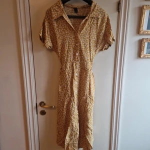 Gul blommig klänning från Shein - Charmig gul klänning från Shein med ett blommigt mönster i vitt. Klänningen har korta ärmar och knappar framtill samt ett knytband i midjan för en fin passform. Perfekt för en solig dag!