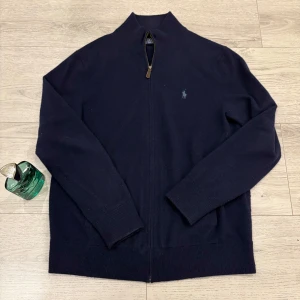 Mörkblå tröja med dragkedja från Ralph Lauren - Säljer en stilren mörkblå tröja från Ralph Lauren med dragkedja och broderad logga på bröstet. Tröjan har långa ärmar och är perfekt för en avslappnad men snygg look. Passar bra till jeans eller chinos. 100% Marinoull. Lite luddig men inget som inte kan fixas med noppbortagare.