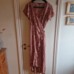 Blommig omlottklänning från Shein - Säljer en söt blommig omlottklänning från Shein i en härlig röd nyans med vita blommor. Klänningen har korta ärmar och en volangdetalj längs nederkanten. Perfekt för sommarens alla tillfällen! 🌸