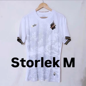 Vit fotbollströja från Nike - Snygg vit fotbollströja från Nike med korta ärmar och stilrena detaljer i svart och guld. Tröjan har ett diskret mönster och klubbmärke på bröstet. Perfekt för fotbollsfans som vill ha något unikt i garderoben.