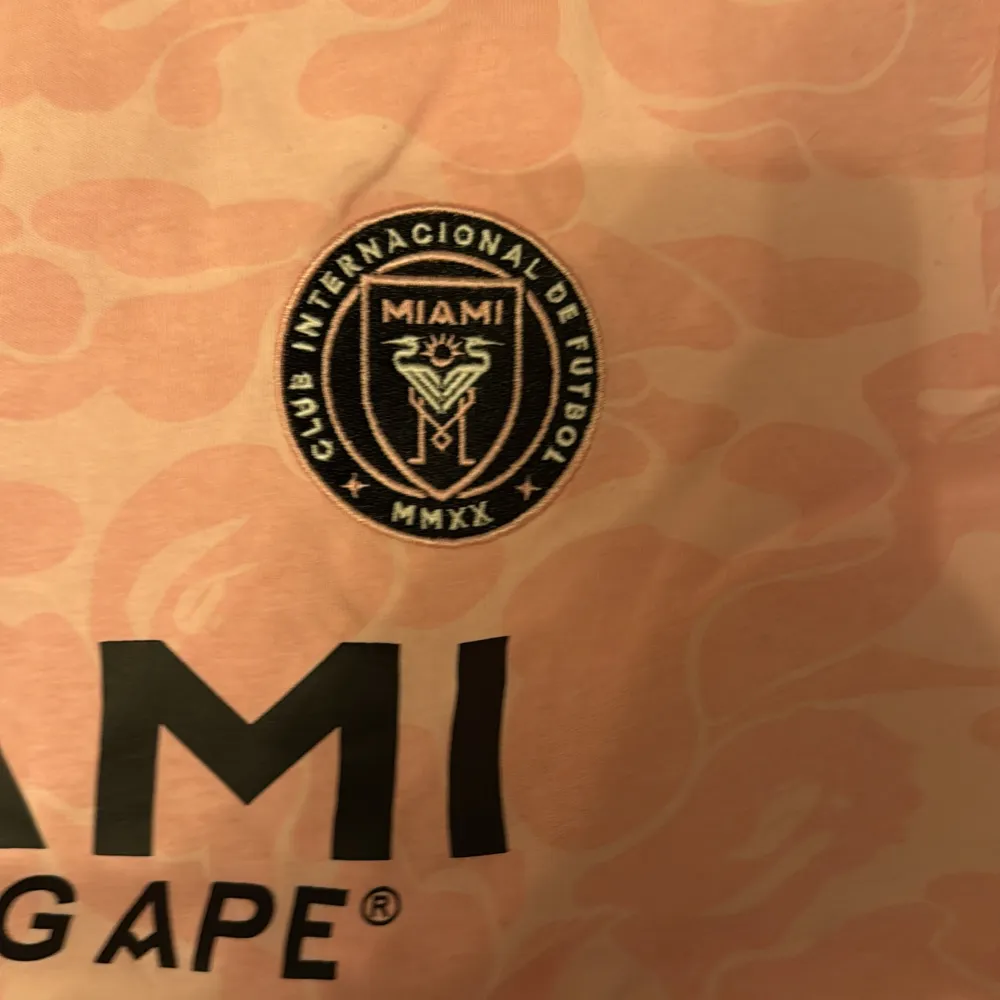 Säljer en rosa fotbollströja från A Bathing Ape i samarbete med Miami. Tröjan har ett unikt mönster och tryck med Miami och A Bathing Ape-loggor. Perfekt för dig som gillar streetwear och fotboll.. T-paidat.