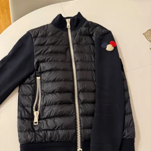 Svart dunjacka med dragkedja - Snygg svart dunjacka från Moncler med quiltad design och dragkedja framtill. Jackan har långa ärmar och är en unik modell med de tre märkerna på ärmen. Perfekt för kyligare dagar till vår och höst!