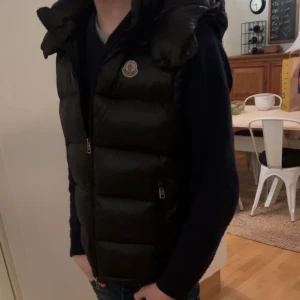 Svart dunväst från Moncler - Snygg svart dunväst från Moncler med dragkedja och broderad logotyp på bröstet. Perfekt för våren och sommaren för den delen med sin snygga och fräscha design och stilrena look.