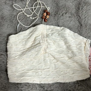 Vit axelbandslös topp från Shein - Säljer en söt vit axelbandslös topp från Shein med dekorativa träpärlor och snörning. Toppen har en vågig textur och är perfekt för varma dagar. Den är enkel att matcha med olika outfits och ger en bohemisk touch.