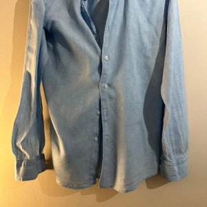 Ljusblå linneskjorta - Snygg ljusblå skjorta med klassisk design och knappar framtill. Perfekt för en stilren look. Skjortan har långa ärmar och är tillverkad i linne material.