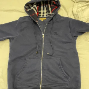 Mörkblå hoodie från Burberry - Säljer en snygg mörkblå hoodie från Burberry med dragkedja och klassiskt rutigt mönster i huvan. Tröjan har en broderad logga på bröstet och praktiska fickor framtill. Perfekt för en stilren och bekväm look. Unik burberry i navy blue. Jätte fin kvalite o d bara höra av sig vid frågor 