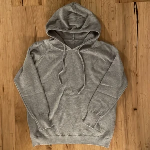 Grå hoodie - Säljer en stilren grå hoodie med justerbar dragsko i huvan. Perfekt för en avslappnad look. Den har långa ärmar och ribbade muddar vid ärmslut och nederkant.