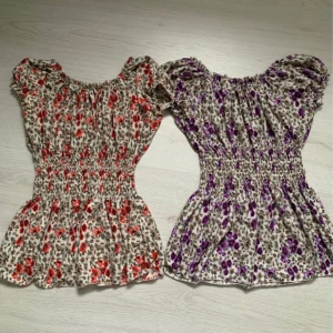 Blommiga blusar  med smockdetaljer - Två blommiga blusar  med smockdetaljer i midjan. Den ena har röda blommor och den andra lila. Båda topparna har korta ärmar och en luftig passform, perfekt för varma dagar.