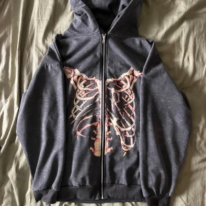 Grå huvtröja med skelettmönster - Cool grå huvtröja med dragkedja och ett unikt skelettmönster på både fram- och baksidan. Perfekt för en edgy look. Tröjan har långa ärmar och en bekväm overzised passform.