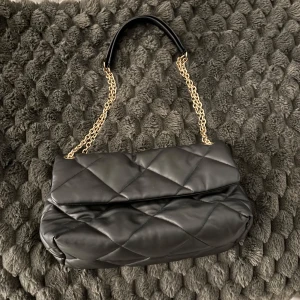 Svart quiltad axelväska med kedja - Snygg svart quiltad axelväska med guldfärgad kedja som axelrem. Väskan har en elegant design och är perfekt för att ge en stilren touch till din outfit.