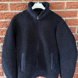 Svart teddyjacka med dragkedja - Mysig svart teddyjacka med dragkedja framtill. Jackan har en hög krage och två fickor på sidorna. Perfekt för kyliga dagar när du vill hålla dig varm och bekväm.