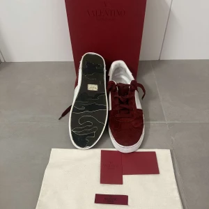Röda sneakers från Valentino - Säljer ett par snygga röda sneakers från Valentino. De är i mocka med vita detaljer och har en klassisk snörning. Skorna är i nyskick och kommer med originalförpackning och dustbag. Perfekta för att ge din outfit en lyxig touch!