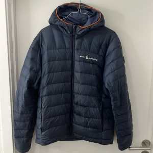 Mörkblå pufferjacka från Sail Racing - Säljer en snygg mörkblå pufferjacka från Sail Racing. Jackan har en dragkedja framtill och en huva med orange detaljer. Spännband både inuti jackan och vid hand,har en logga på bröstet och en patch på ärmen. Skicket 6-7/10, skulle behöva liten tvätt.