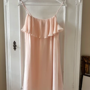 Ljusrosa axelbandslös klänning - Säljer en superfin ljusrosa klänning med smala flätade axelband. Den har en volangdetalj upptill som ger en romantisk touch. Perfekt för sommarens fester eller en mysig midsommar. Klänningen är i ett lätt och luftigt material