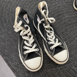 Svarta Converse All Star sneakers - Säljer ett par klassiska svarta Converse All Star sneakers i storlek dam 36. De har den ikoniska vita tåhättan och snörning. Skorna är unisex och tillverkade i textil med en man-made sula. Perfekta för både vardag och fest!