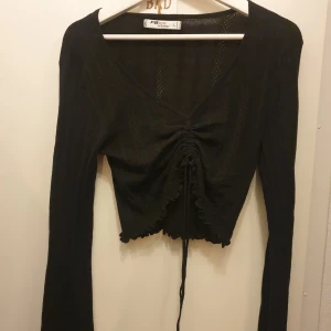 Svart ribbad topp från F&F - Säljer en snygg svart ribbad topp från F&F. Den har långa vida ärmar och en v-ringad design med knytning framtill som ger en cool look. Perfekt för både vardag och fest! Passar allt från S-L beroende hur man vill att den ska sitta, den är stretchig.