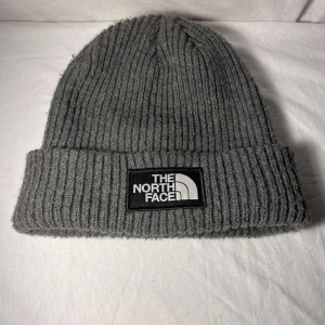 Grå ribbad mössa från The North Face - Säljer en stilren grå ribbad mössa från The North Face. Mössan har en uppvikt kant och ett tydligt märke framtill. Perfekt för kyliga dagar.