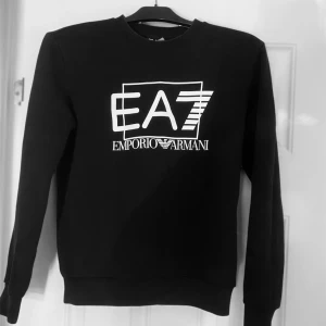 Svart tröja från Emporio Armani - Snygg svart tröja från Emporio Armani med det ikoniska EA7-logotypen i vitt på framsidan. Tröjan har långa ärmar och en klassisk rund halsringning. Perfekt för en stilren och modern look. Priset kan såklart diskuteras 