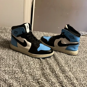 Air Jordan 1  - Snygga Nike Air Jordans i en fräsch kombination av blått, svart och vitt. Skorna har en hög design med klassisk snörning och den ikoniska Nike-loggan på sidan.