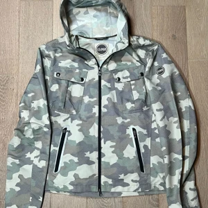 Camouflage vindjacka från Colmar - Säljer en snygg camouflage vindjacka från Colmar. Jackan har en huva och dragkedja framtill, samt praktiska fickor med dragkedjor. Perfekt för utomhusaktiviteter och har en cool look med sitt mönster. Storleken M jätte bra sick
