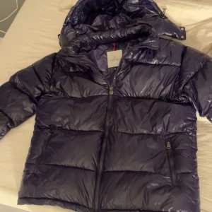 Mörkblå dunjacka från Moncler - Säljer en snygg svart dunjacka från Moncler. Jackan har en glansig finish och är utrustad med en dragkedja framtill. Den har en huva och en ficka med knappdetalj på ärmen. Perfekt för kyliga dagar!