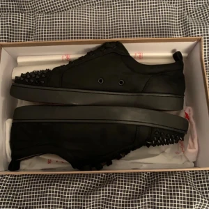 Svarta sneakers med nitar från Christian Louboutin - Snygga svarta sneakers från Christian Louboutin med nitar på ovansidan. Skorna har en klassisk röd sula och är tillverkade i ett stilrent svart material. Perfekta för att ge en edgy touch till din outfit.