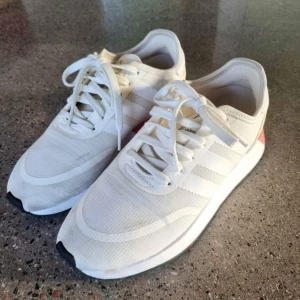 Vita sneakers från Adidas - Snygga vita sneakers från Adidas med klassiska tre ränder på sidan. Skorna har en vit ovandel med snörning och en kontrasterande röd detalj på hälen. 