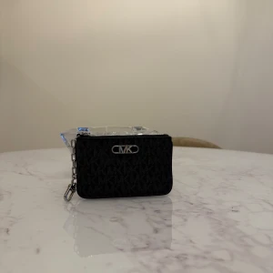 Svart plånbok från Michael Kors - Snygg och kompakt svart plånbok från Michael Kors med ett diskret mönster och en praktisk dragkedja. Perfekt för att hålla ordning på kort och småsaker. Den har en metallkedja för extra säkerhet och stil.