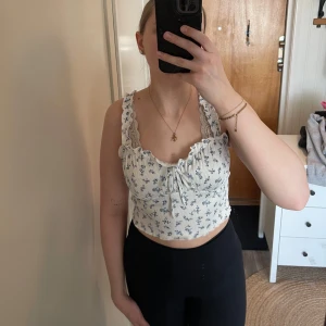 Blommig vit topp från Shein - Söt vit topp med blå blommor från Shein. Toppen har volangdetaljer och knytning framtill. Den är ärmlös med smala axelband och har en ribbad struktur.