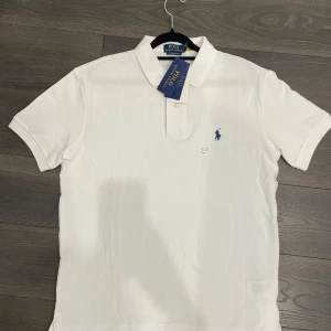Vit pikétröja från Ralph Lauren - Säljer en klassisk vit pikétröja från Ralph Lauren i slim fit. Tröjan har en broderad blå logotyp på bröstet och korta ärmar. Perfekt för en stilren look.