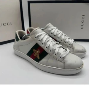 Vita sneakers från Gucci med broderad detalj - Snygga vita sneakers från Gucci med klassiska gröna och röda ränder samt en broderad guldfärgad bi. Skorna har snörning och en stilren design som passar perfekt för en trendig look. Endast påse