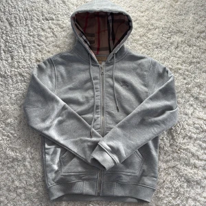 Grå hoodie från Burberry - Säljer en stilren grå hoodie från Burberry med dragkedja och klassiskt rutigt mönster i huvan. Tröjan har en broderad logga på bröstet och är tillverkad i mjukt material för extra komfort. Perfekt för en avslappnad och stilig look. Hoodien är helt ny, använd en gång och tags fås med om det önskas. Säljer den då den var lite för liten för mig och passar mer L än M.